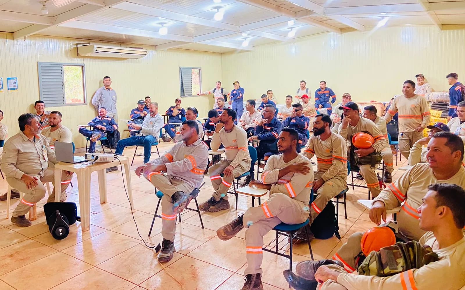 Turma de operadores e equipe de campo do Grupo FX Minas, de uniforme com faixa refletiva, num treinamento em sala, etapa obrigatória antes de assumir a função de operador de máquina pesada